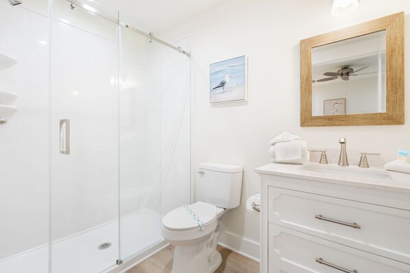 A modern bathroom with a glass shower enclosure, a white toilet, a vanity with a mirror, and a ceiling fan. - Luxury Sandpiper Loft in WaterWay Village Entertainment District (Gulf Shores)
