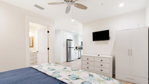 A bedroom with a bed, a wardrobe, a television, and a kitchen in the background.