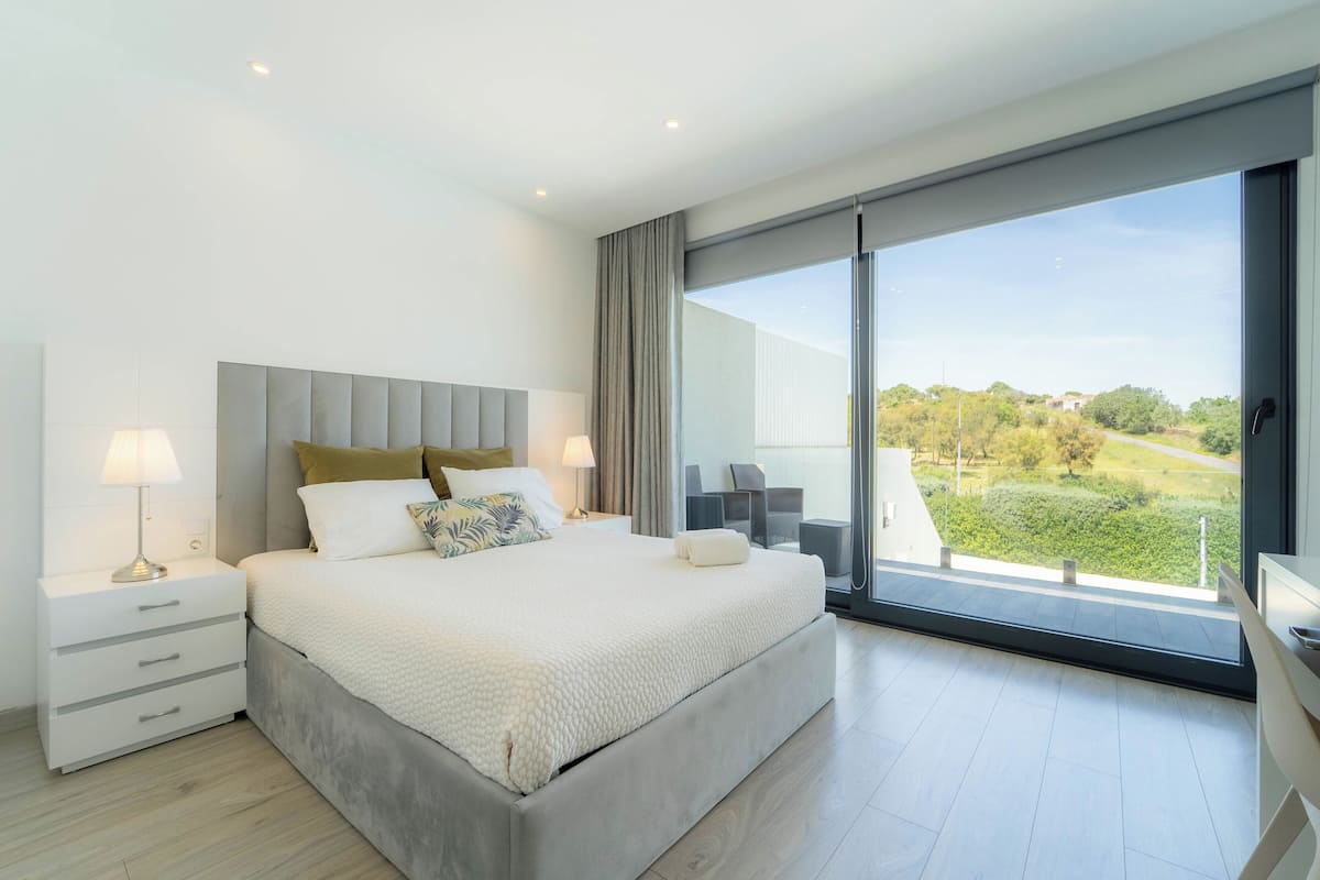 A modern bedroom with a large bed, bedside tables, a view of greenery through a sliding glass door, and a balcony with outdoor furniture.