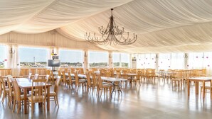 Un salón de banquetes con varias mesas y sillas, una araña de cristal y amplias ventanas con vistas a la ciudad.
