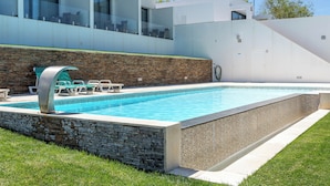 Una piscina moderna con pared de piedra y barandillas de vidrio, rodeada de un césped bien cuidado.