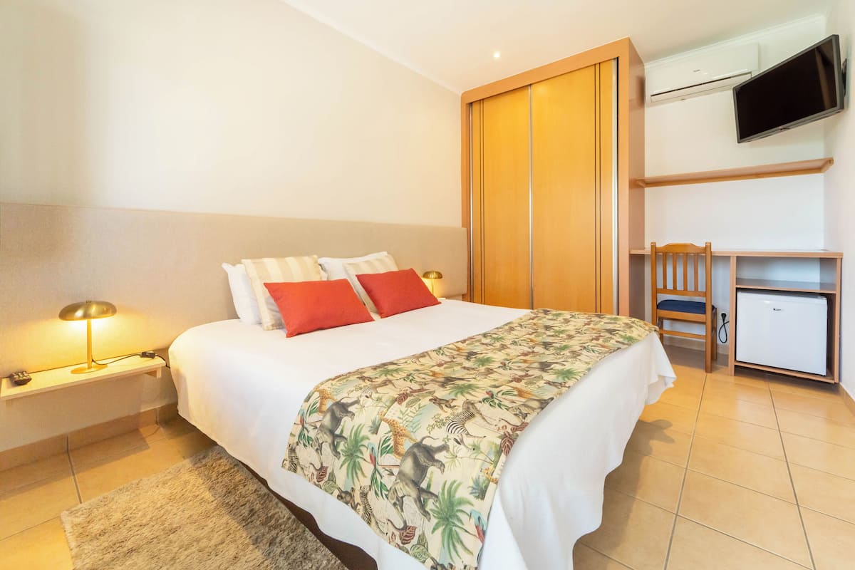 A hotel room with a bed, bedside lamps, a desk, a chair, a TV, and a mini-fridge.