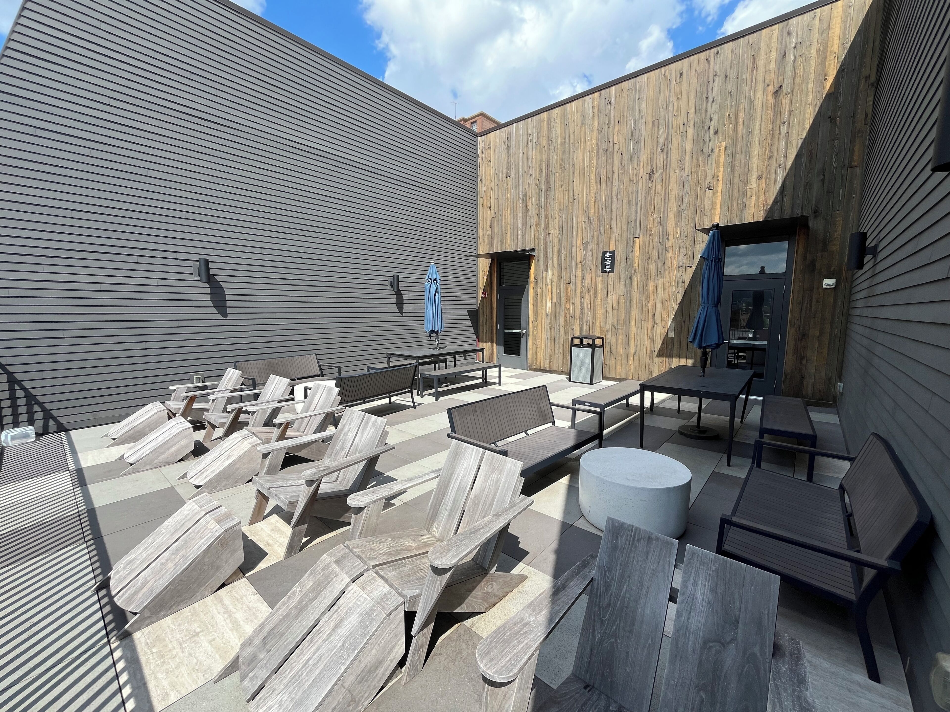 An outdoor seating area with wooden and metal furniture, a blue umbrella, and a stone planter.