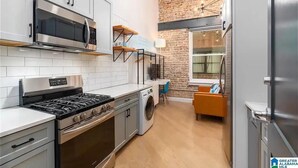Une cuisine compacte dotée d’appareils électroménagers modernes, d’un lave-linge et d’un coin repas agrémenté d’un mur en briques et d’une fenêtre.