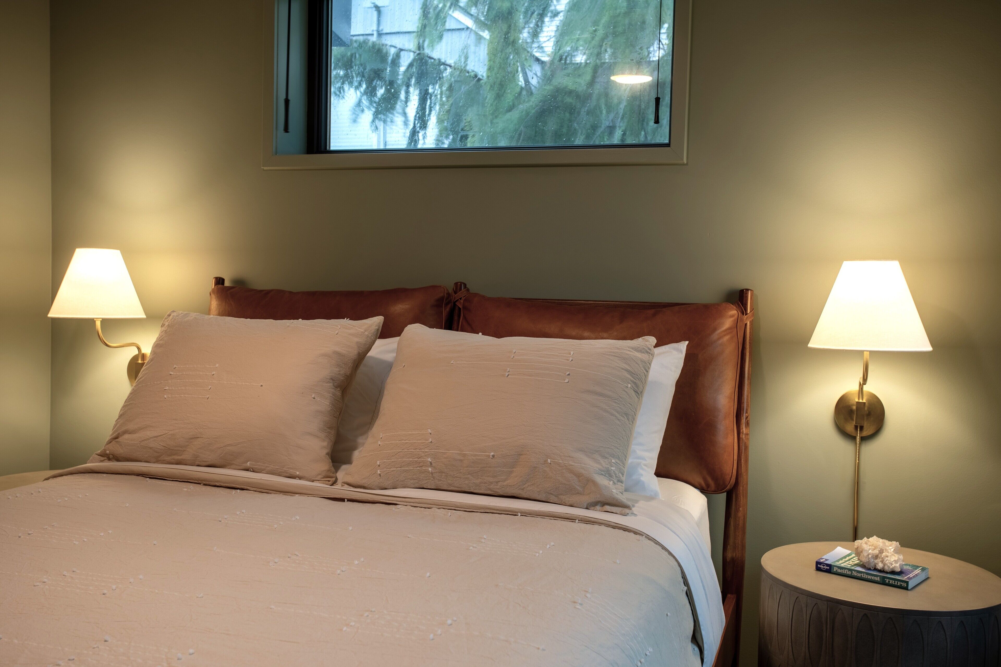 A neatly made bed with two pillows, a round bedside table with a lamp, and a window with a view of trees.