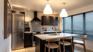 A modern kitchen with a stainless steel refrigerator, a built-in oven, a gas stove, and a large island with bar stools. There are pendant lights above the island and a window with blinds to the right. - Chalet Suite in Downtown Juneau (Juneau)