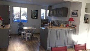 A compact kitchen with grey cabinets, a small dining area with a round table, and a window with a view of a bridge. - Maison Proche Plage Pour 8 Avec Jardin et Internet (ARZON)