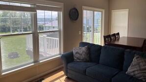 A living room with a blue couch, large windows, and a view of a backyard. - Bayview at Three Schooners Landing (Sodus Point)