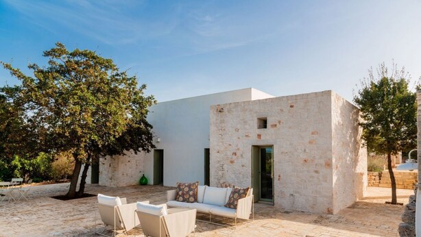 Un espacio moderno al aire libre con un sofá y sillas blancos, un muro de piedra y un árbol.
