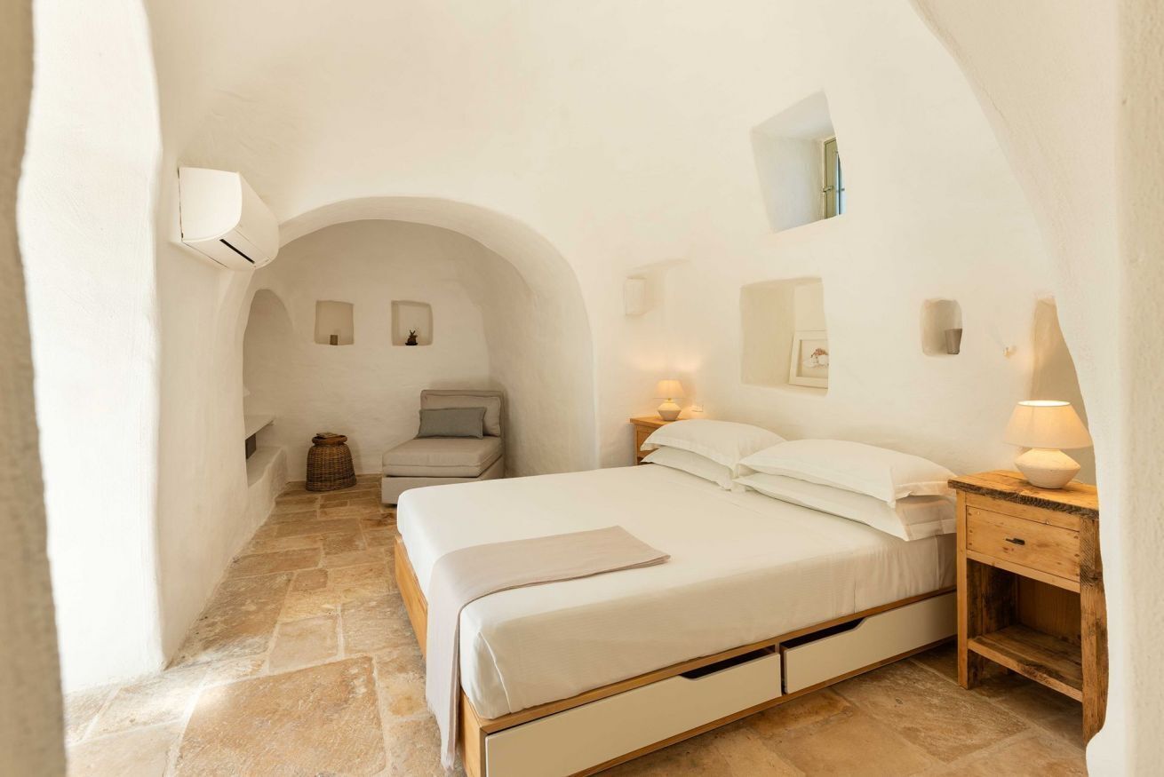 A bedroom with a single bed, a nightstand, a lamp, and a small table with a candle. There is a chair and a small table with a candle in the corner. The room has white walls and a stone floor.