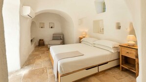 Un dormitorio con una cama individual, una mesita de noche, una lámpara y una pequeña mesa con una vela. Se aprecia una silla y una pequeña mesa con una vela en la esquina. Las paredes son blancas y el suelo, de piedra.