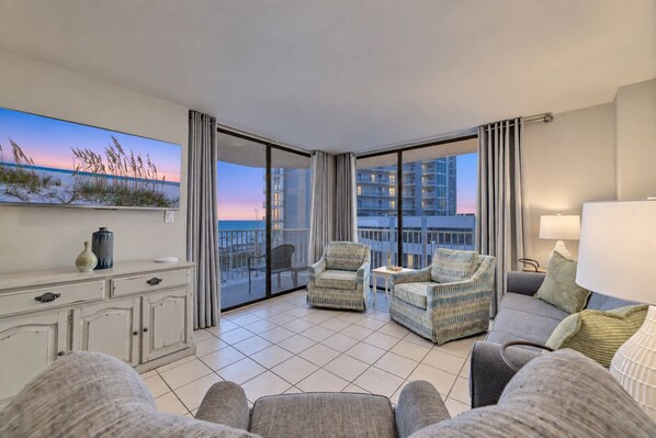 A spacious balcony with a view of the sea and buildings, furnished with a sofa, chairs, and a small table.