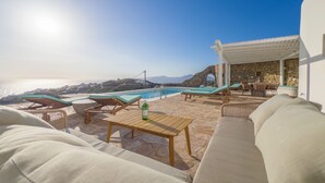 A sunlit outdoor seating area with lounge chairs and a pool, overlooking a coastal landscape. - Villa Prive 77 Mykonos (Mykonos)