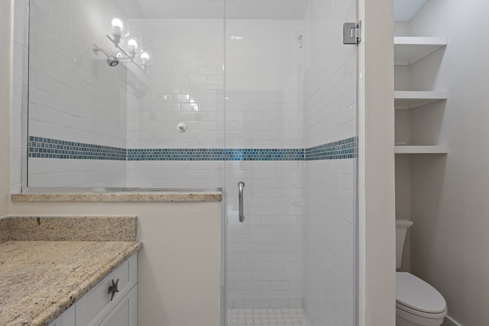 A modern bathroom with a glass-enclosed shower, white tiled walls, a granite countertop, and a toilet visible through an open door.