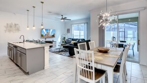 A modern kitchen with a central island, dining area with white chairs, and a large window providing natural light. - Modern 4BR Home with Private Pool near Disney (Kissimmee)