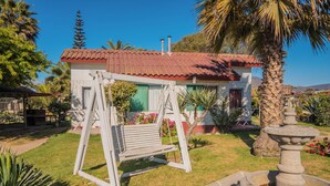 Una casa con techo de tejas rojas, un columpio, una fuente y un jardín bien cuidado con palmeras y plantas florecientes.