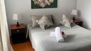 Un dormitorio ordenado con una cama, dos mesitas de noche, una lámpara, un almohadón y un cuadro colgado en la pared.