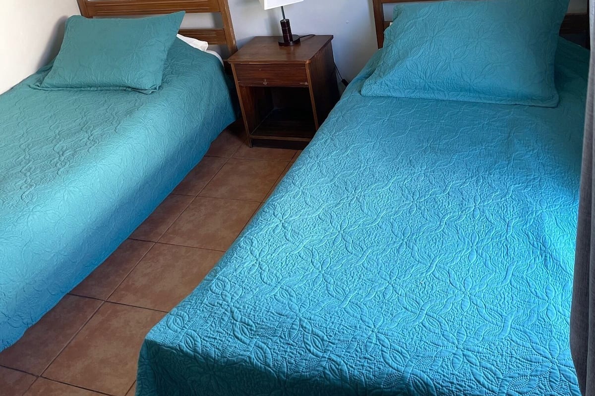 Una habitación con dos camas individuales, cada una con cubrecama y almohadas color turquesa. Hay una mesita de noche de madera con una lámpara entre las camas. Las paredes son blancas con un diseño de ladrillo, y hay dos pequeños cuadros enmarcados colgados en la pared.