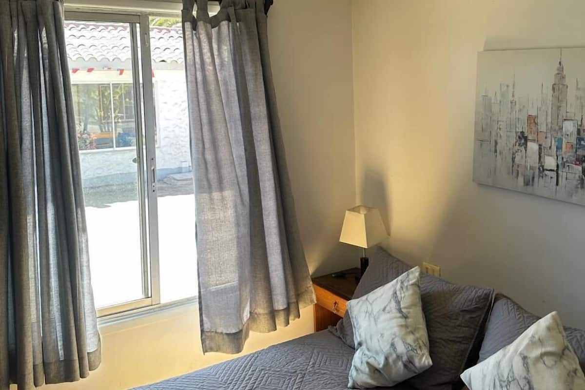 Un dormitorio con cama, mesita de noche, lámpara y ventana con cortinas.