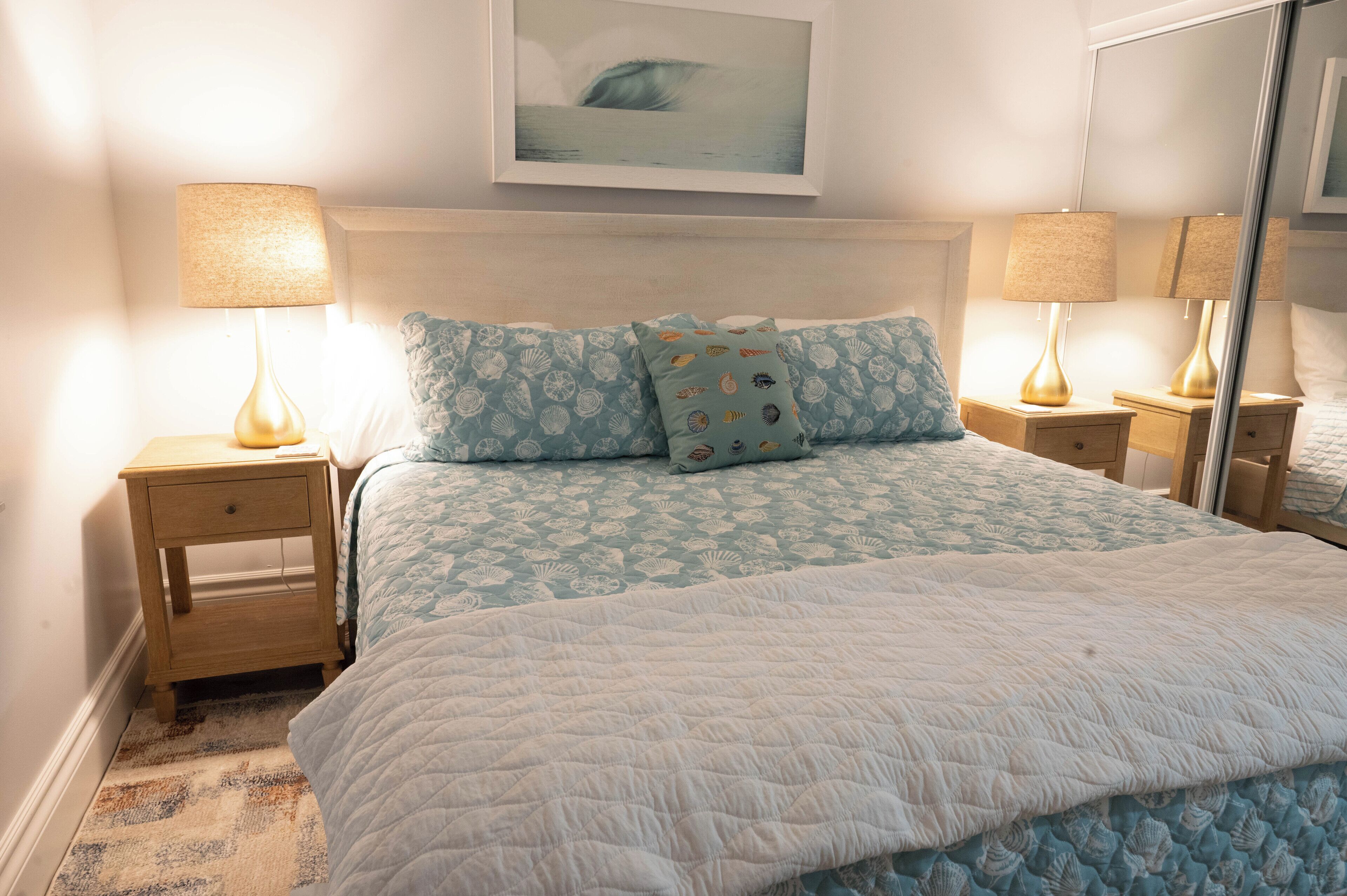 Una cama bien hecha con una cubierta blanca acolchada, dos lámparas iguales sobre mesitas de noche de madera, y un cuadro enmarcado sobre la cama.