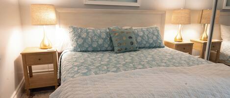Una cama bien hecha con una cubierta blanca acolchada, dos lámparas iguales sobre mesitas de noche de madera, y un cuadro enmarcado sobre la cama.