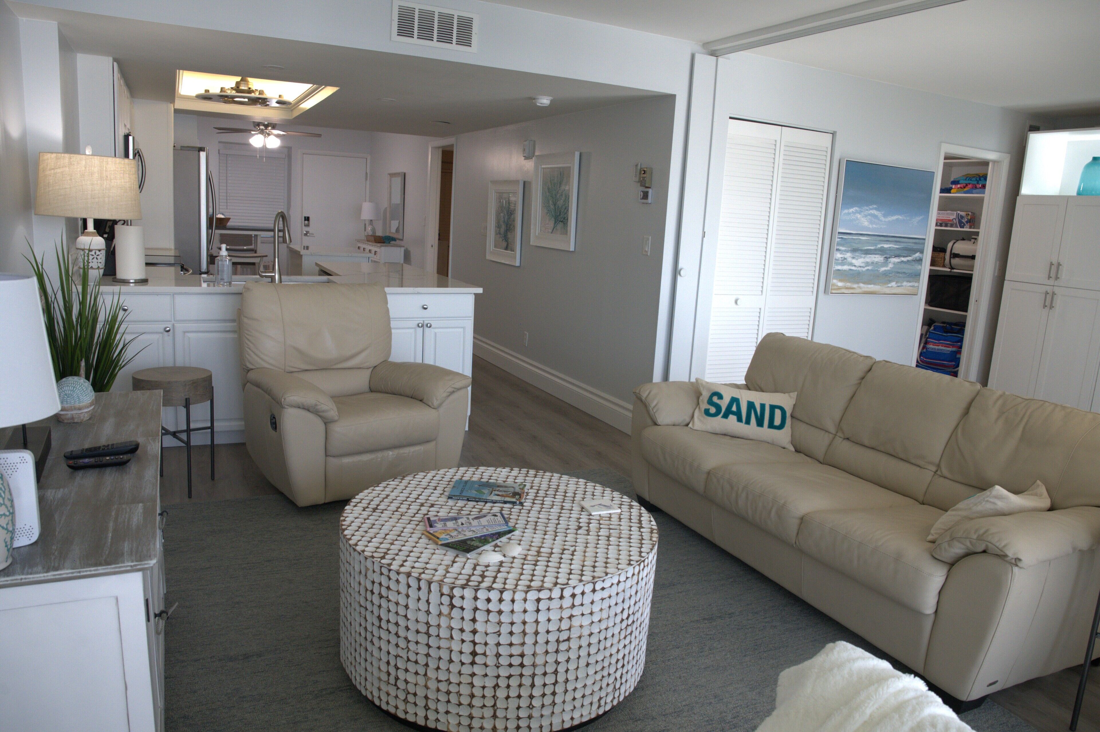 A living room with a beige sofa, a round ottoman, a beige armchair, a small round table, and a kitchen area in the background.