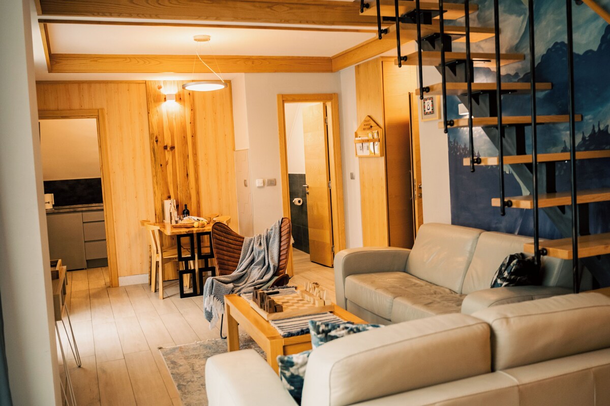 Un salon moderne comprenant un canapé, une table basse et un coin repas avec une table et des chaises. On y trouve un escalier avec une rampe en bois et un tableau accroché au mur.