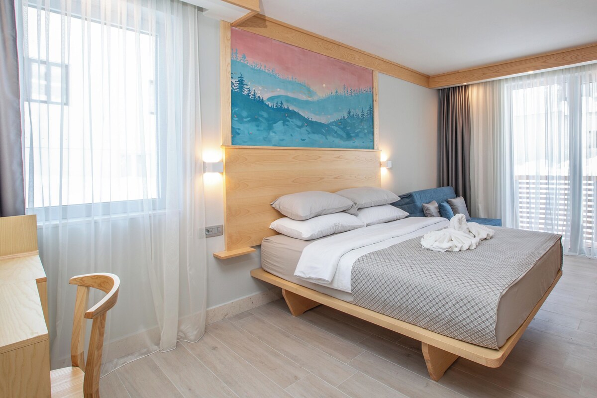 A bedroom with a bed, a chair, a painting, and a logo on the floor.