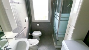 Un baño compacto con lavamanos, dos inodoros, cabina de ducha de cristal y lavadora integrada.