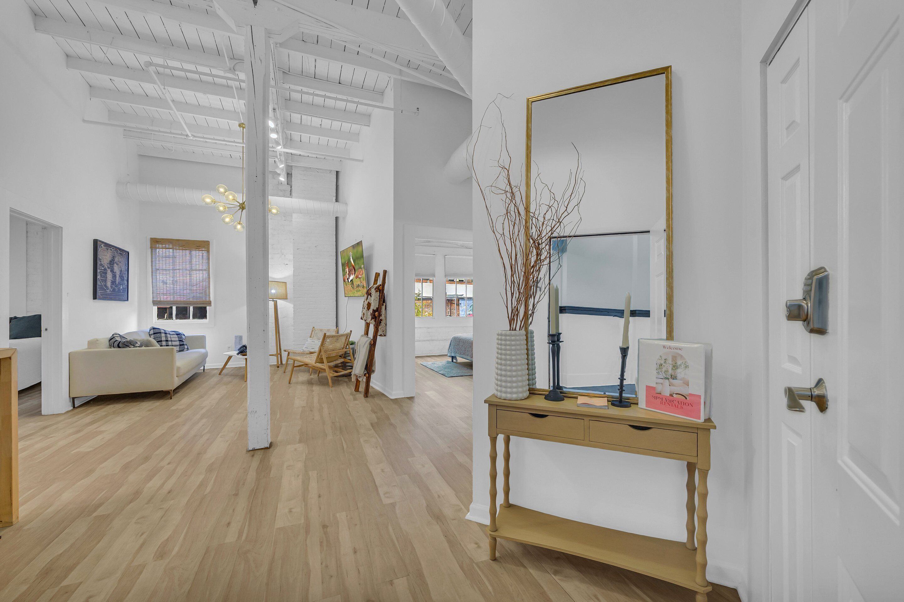 Una habitación amplia con piso de madera, un sofá blanco, una consola de madera con espejo y una puerta con pomo de latón.