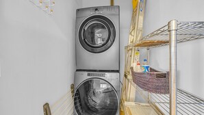A compact laundry area with two stacked washing machines and a metal shelf with a basket and bottles. - The Dawg Walk Downtown (Athens)