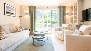 Un salon moderne avec un canapé, un fauteuil et une table basse. Il y a une grande fenêtre avec des rideaux, un téléviseur à écran plat fixé au mur et un tableau encadré accroché au mur.