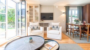 Un salon moderne avec un grand téléviseur, des fauteuils blancs, une table à manger avec des chaises et une table basse en verre.