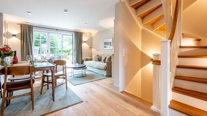 A modern living room with a wooden staircase, a dining area with chairs and a table, and a sofa. - Landhaus Senwai 3 - Neubau. Stadtnah. Wellness (Sylt)