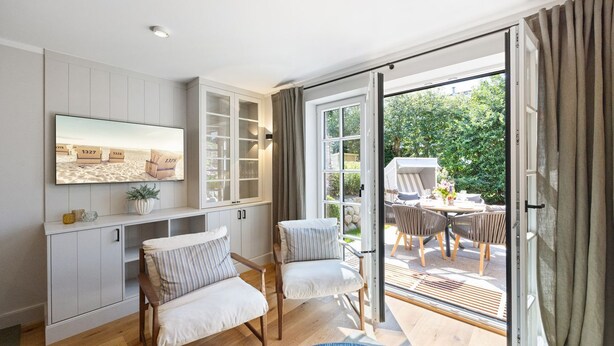 A modern living room with a TV, built-in cabinets, and a sliding glass door leading to an outdoor patio. - Landhaus Senwai 3 - Neubau. Stadtnah. Wellness (Sylt)