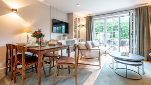 A modern living room with a dining area, a TV mounted on the wall, and a large sliding glass door leading to an outdoor area. - Landhaus Senwai 3 - Neubau. Stadtnah. Wellness (Sylt)
