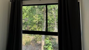 A room with a bed, a window with black curtains, and a view of greenery outside. - Modern Family - sized  2 bedroom 2 bath condo just  10 min walk to the beach (Puntarenas Province)