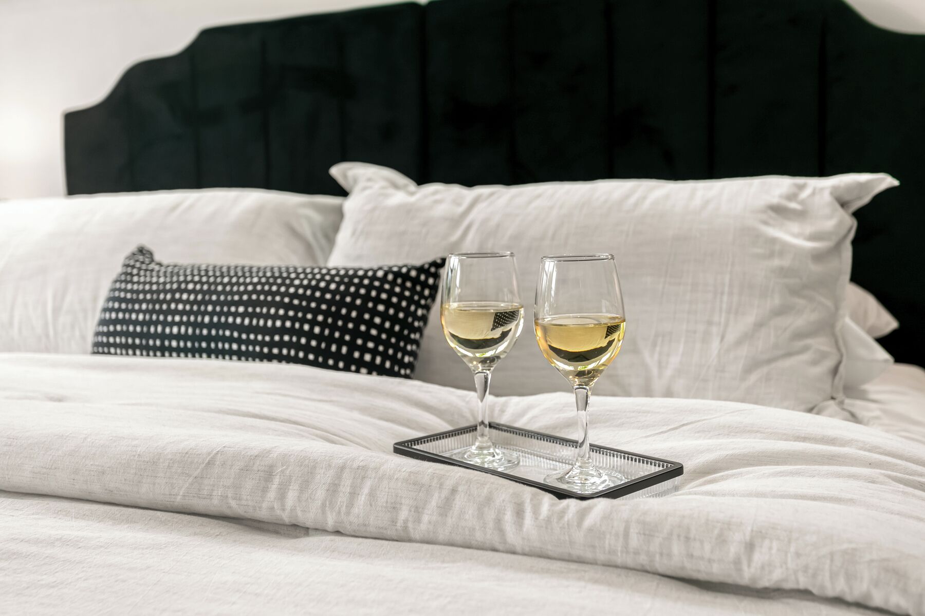 Un letto con due bicchieri di vino bianco su un vassoio, una testiera nera e un cuscino bianco a pois.
