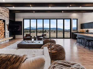 Ein modernes Wohnzimmer mit Kamin aus Stein, Ledermöbeln, einem großen Fenster mit Blick auf die Berge und einer Küchenzeile.