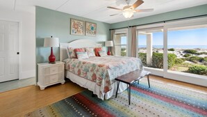 Une chambre à coucher comprenant un lit, des tables de chevet, un banc, un ventilateur de plafond et une grande fenêtre donnant sur la plage.