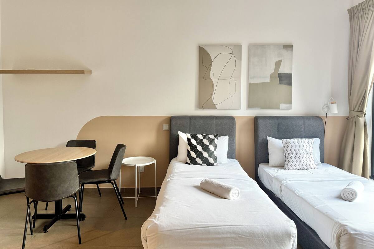 Habitación de hotel con dos camas individuales, una mesita redonda pequeña, una silla y una lámpara fijada a la pared.