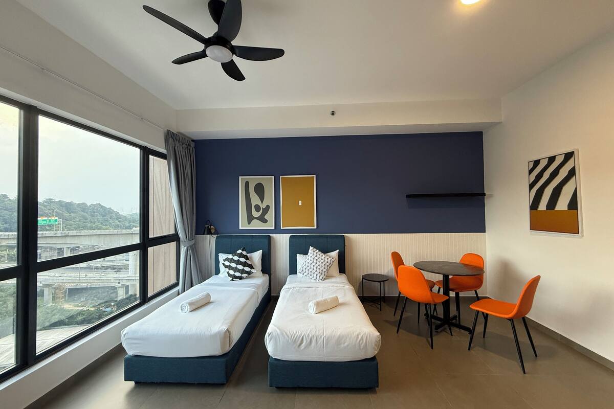 Habitación de hotel con dos camas, ventilador de techo, una mesa de comedor con sillas y un ventanal con vistas a la ciudad.