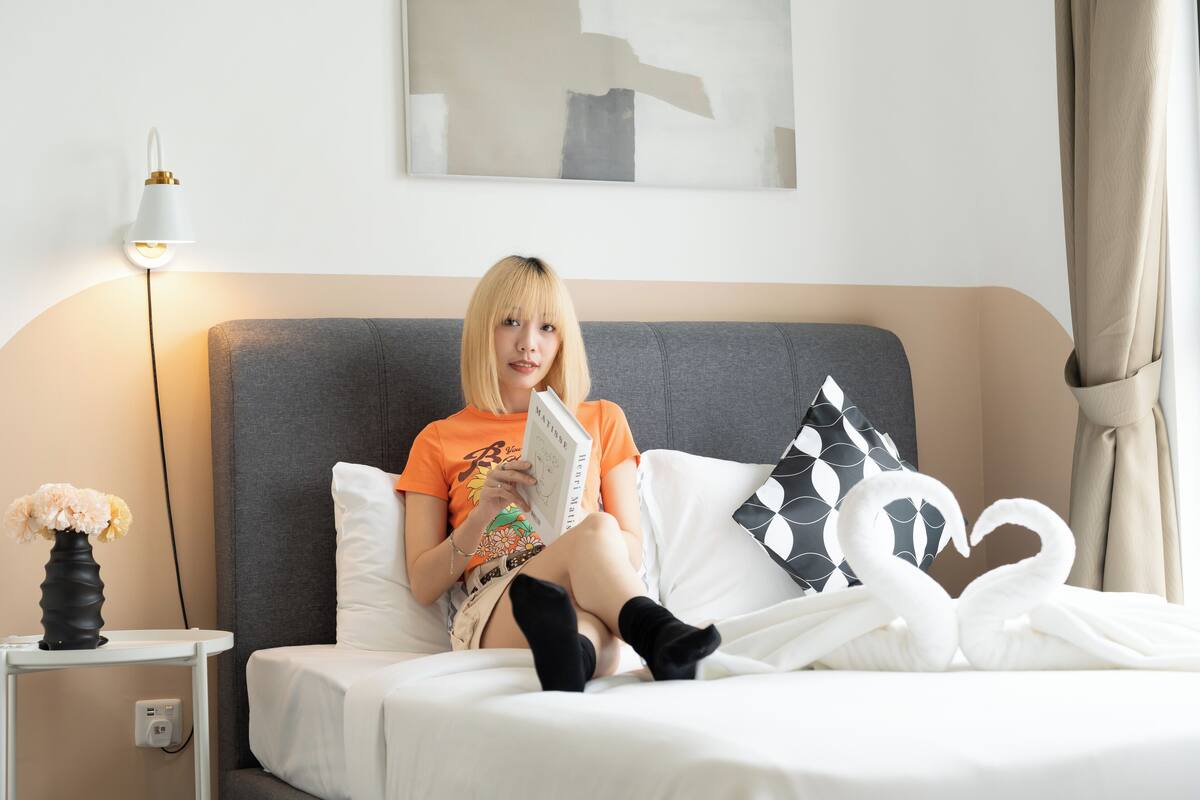 Una persona sentada en una cama con un libro, un jarrón de flores y una lámpara.