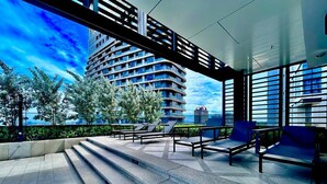 Un área de estar moderna al aire libre con escalones que conducen a un edificio, rodeada de follaje y un cielo despejado.