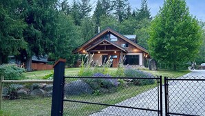 A wooden cabin with a garage, surrounded by lush greenery and tall trees. - "Sleeps 15: Dog-Friendly Rustic Haven on Cortes Island" Large grass yard (Mansons Landing)