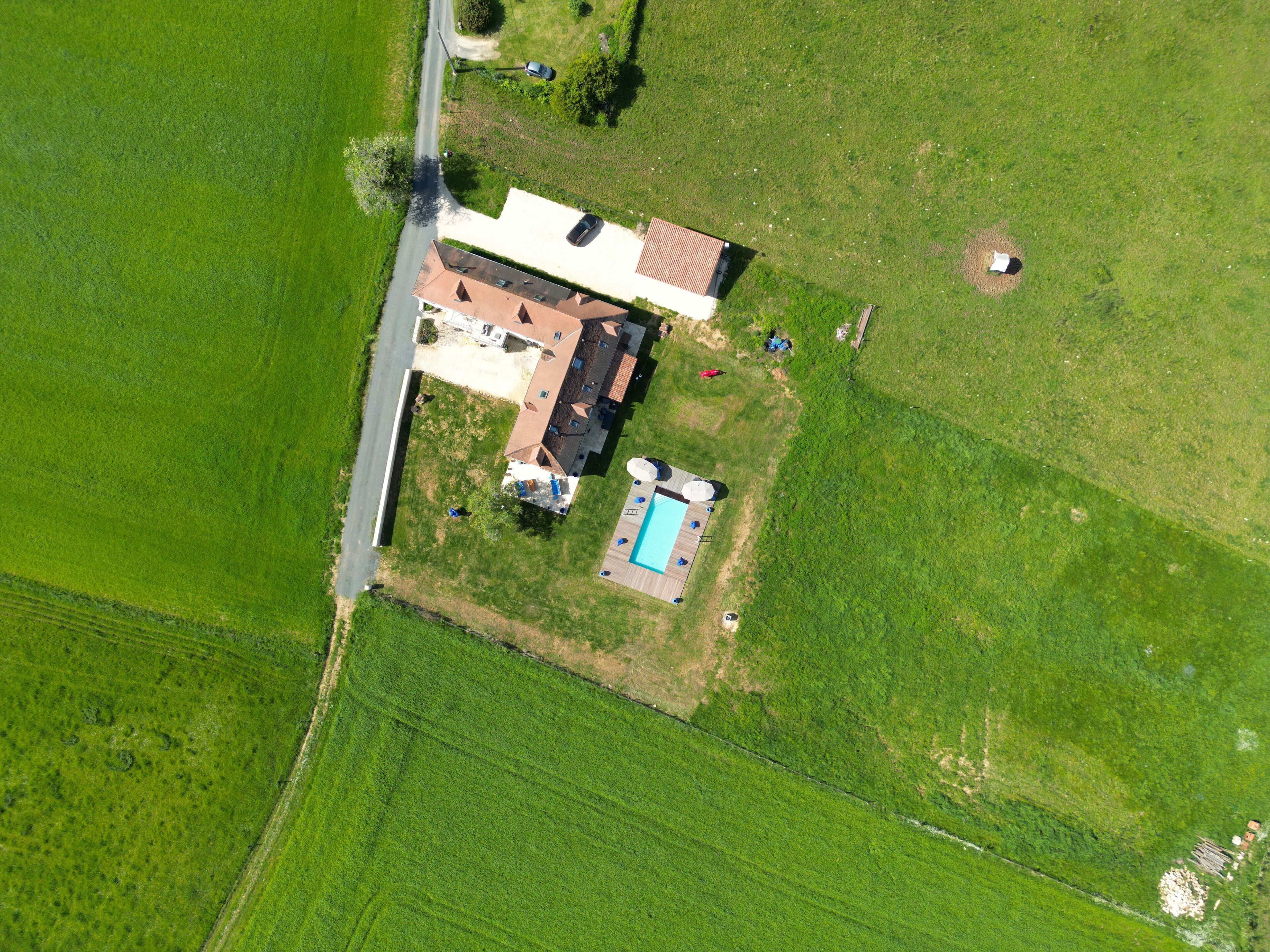 An aerial view of a house with a swimming pool, surrounded by green fields.
