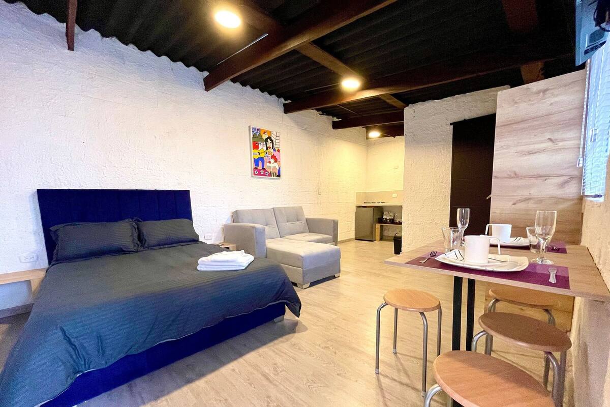 A bedroom with a bed, a sofa, a dining table with chairs, and a kitchen area.