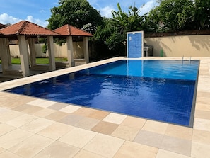A rectangular swimming pool with a tiled deck, a covered area with a red roof, and a blue fence. - Hosts BR - Apartamentos funcionais (Fortaleza)