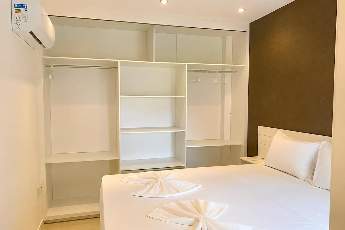 Quarto de hotel com uma cama grande branca, um guarda-roupa com várias prateleiras e uma mesa de cabeceira.
