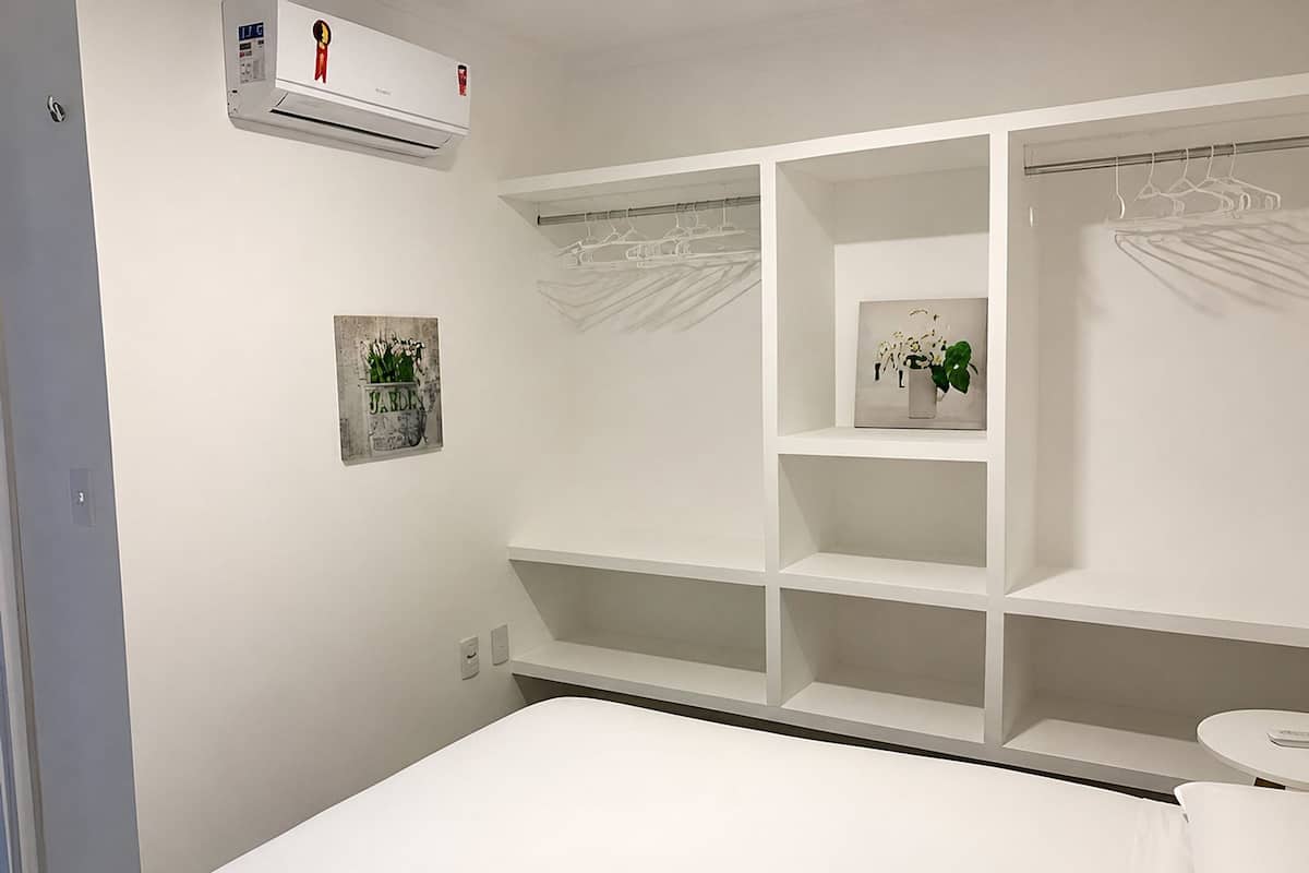 Quarto com cama, guarda-roupa branco com espaço para pendurar roupas e duas prateleiras fixadas na parede com plantas em vasos.